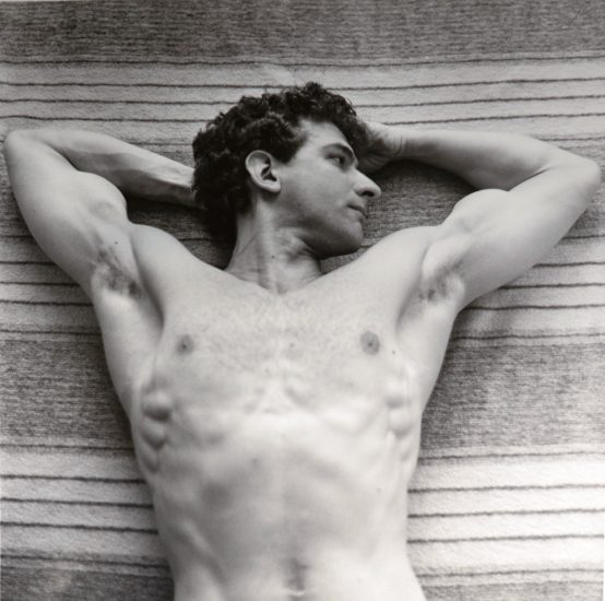 This black and white photograph shows a shirtless man lying on a towel with his arms behind his head.