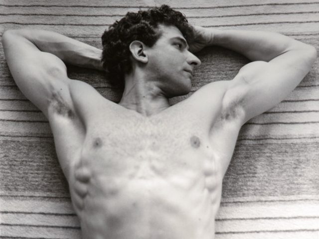 This black and white photograph shows a shirtless man lying on a towel with his arms behind his head.