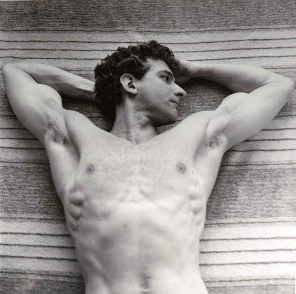 This black and white photograph shows a shirtless man lying on a towel with his arms behind his head.