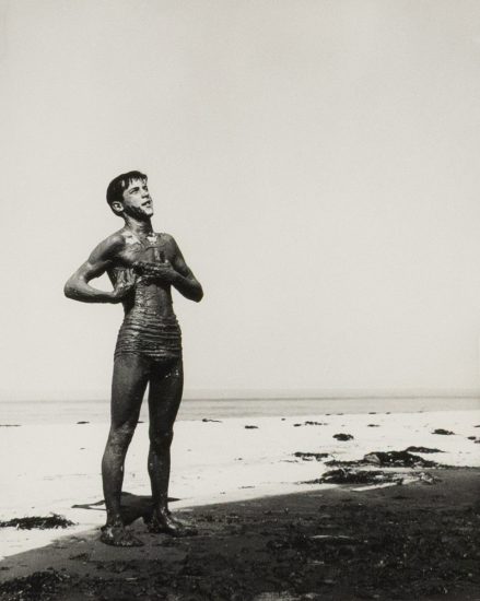 This black and white photograph shows a naked young man standing on a beach. He is covered in mud.