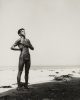 This black and white photograph shows a naked young man standing on a beach. He is covered in mud.