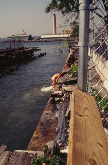This color photograph shows a naked man climbing out of a river.