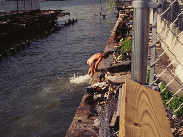 This color photograph shows a naked man climbing out of a river.