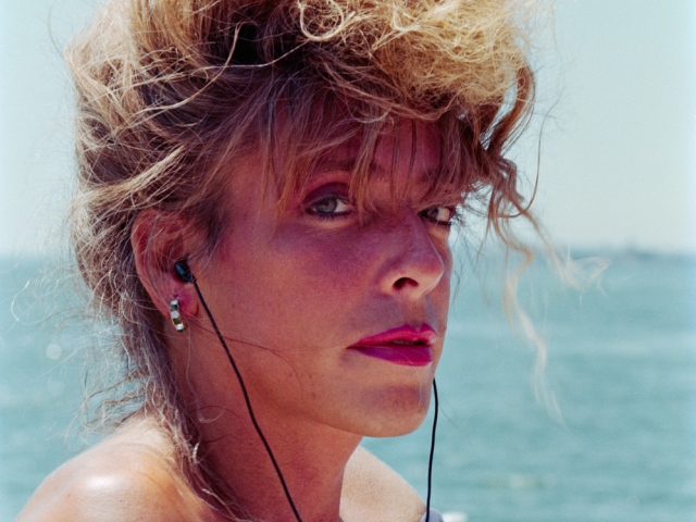 This color photograph is a portrait of a woman from the chest up. She has red hair piled on her head and wears ear buds.