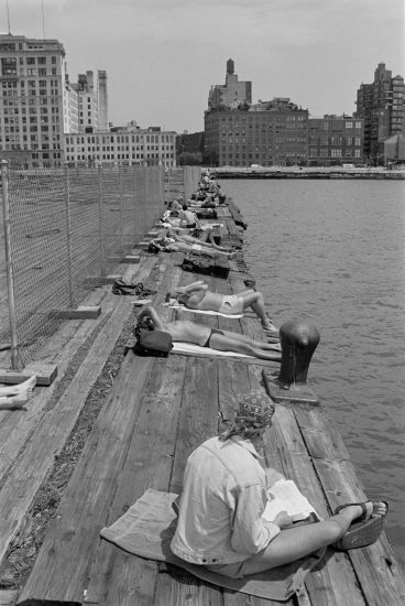 This black and white photograph shows a wooden pier overlooking New York City. A line of men sits on the pier.