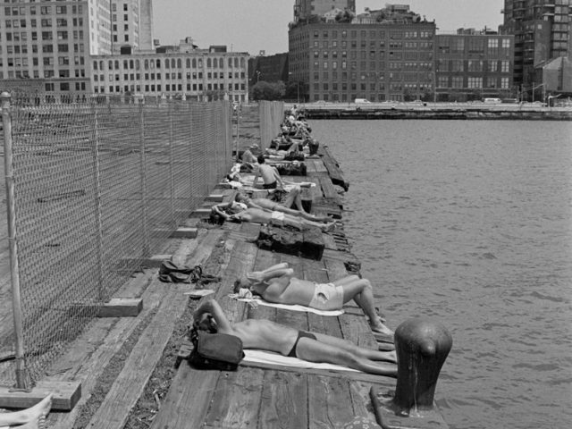 This black and white photograph shows a wooden pier overlooking New York City. A line of men sits on the pier.