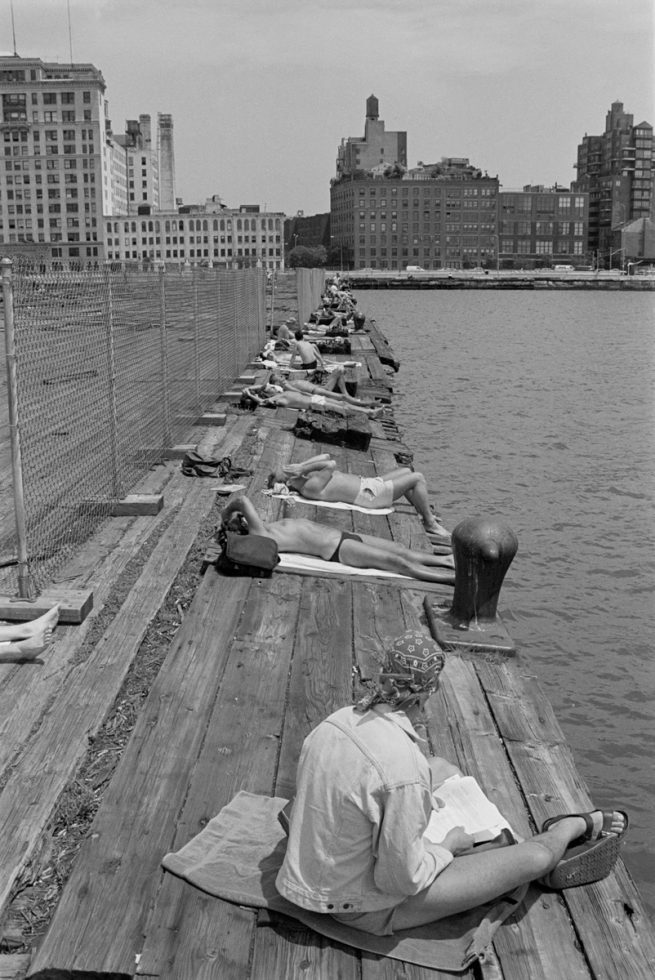 This black and white photograph shows a wooden pier overlooking New York City. A line of men sits on the pier.