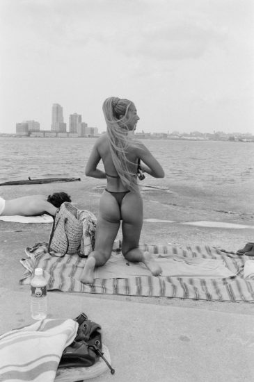 This black and white photograph shows the back of a woman kneeling on a pier overlooking the water.