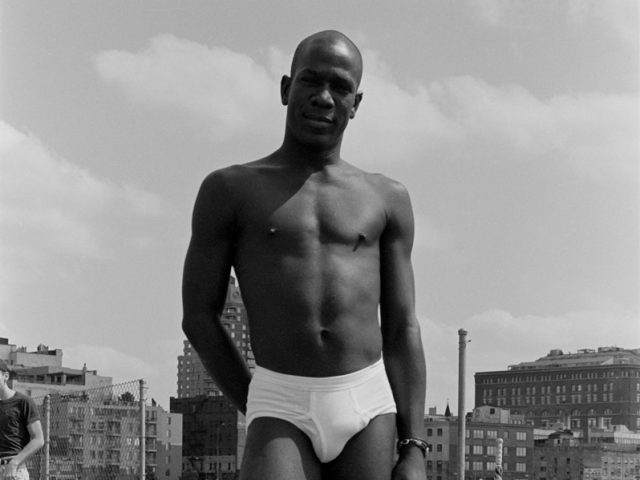 This black and white photograph depicts a shirtless man standing on the pier with one foot lifted a bit off the plank.