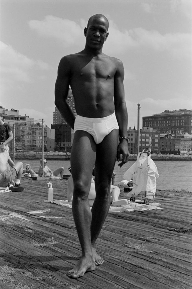 This black and white photograph depicts a shirtless man standing on the pier with one foot lifted a bit off the plank.