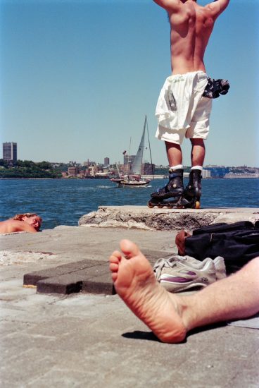 This color photograph shows a landscape of a pier, with a bare foot in the foreground, and a shirtless man with his hands above his head in the background.