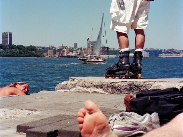 This color photograph shows a landscape of a pier, with a bare foot in the foreground, and a shirtless man with his hands above his head in the background.