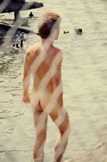 This color photograph shows a naked man seen from behind a chain link fence. He overlooks the water.