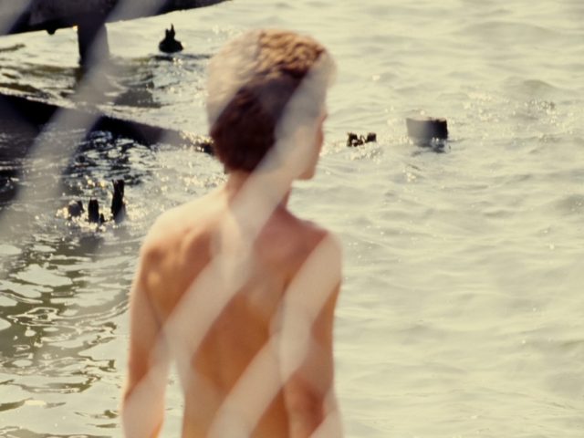 This color photograph shows a naked man seen from behind a chain link fence. He overlooks the water.
