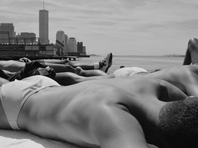 This black and white photograph overlooks the bare back of a man lying on a pier overlooking skyscrapers rising from the water.