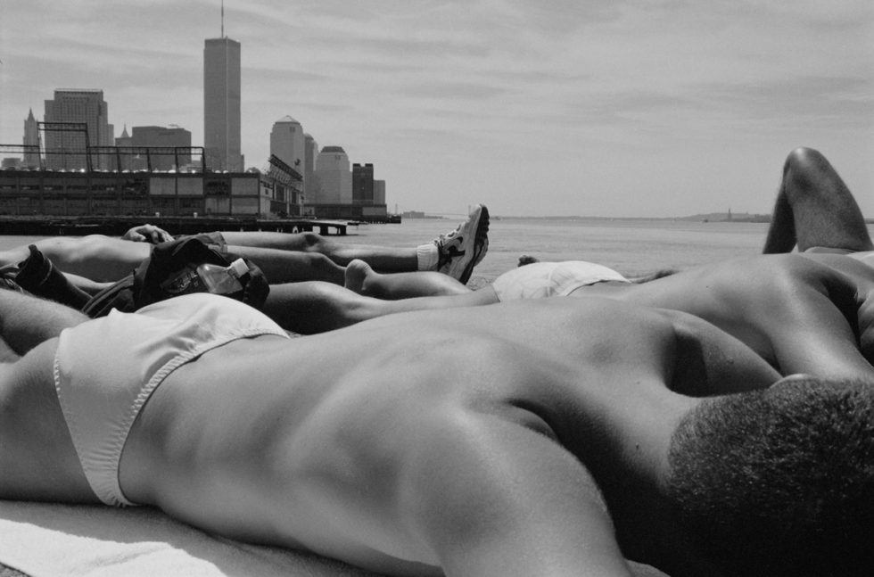 This black and white photograph overlooks the bare back of a man lying on a pier overlooking skyscrapers rising from the water.