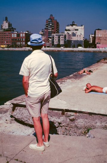 This color photograph shows the back of a tourist wearing a green shirt. He stands on a concrete pier overlooking the water.