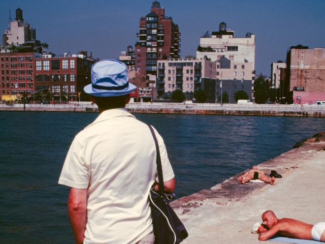 This color photograph shows the back of a tourist wearing a green shirt. He stands on a concrete pier overlooking the water.
