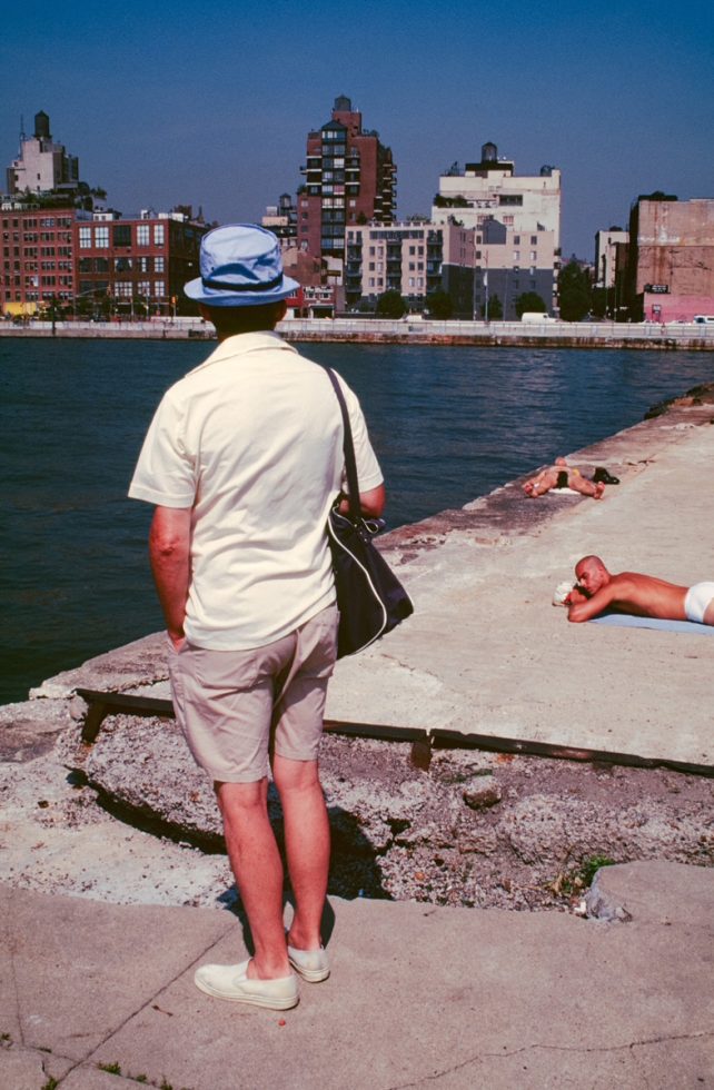 This color photograph shows the back of a tourist wearing a green shirt. He stands on a concrete pier overlooking the water.
