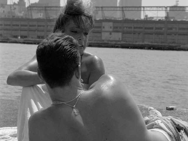 This black and white photograph shows the back of a shirtless man overlooking the New York skyline on a pier.