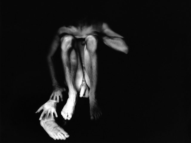 This black and white photograph shows a glowing figure crouching on the ground against a black background.