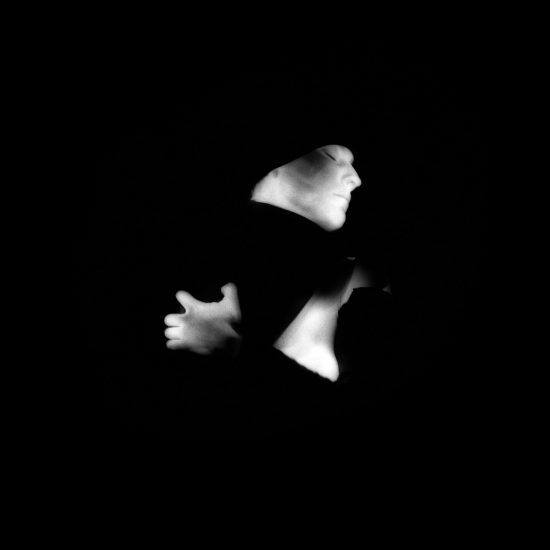 This black and white photograph shows a woman with her arms wrapped around herself. She is seen from the side, and her skin glows white against a black background.