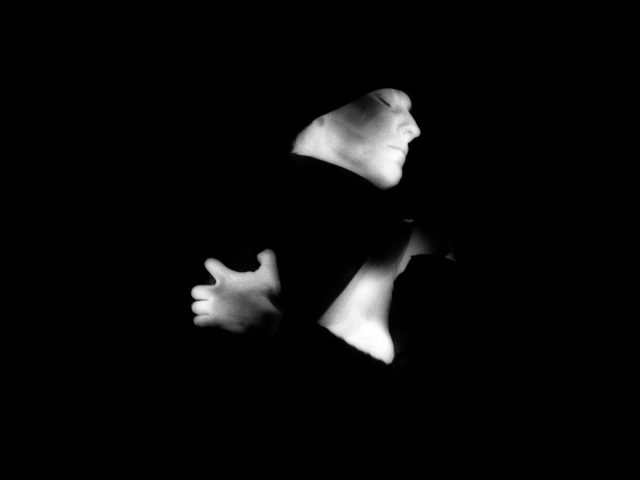 This black and white photograph shows a woman with her arms wrapped around herself. She is seen from the side, and her skin glows white against a black background.
