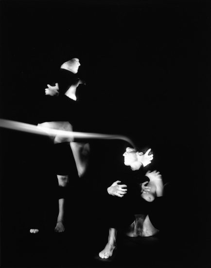 This black and white photograph shows multiple glowing white figures, set against a black background. Their arms are wrapped around themselves and their bodies are blurred with motion.