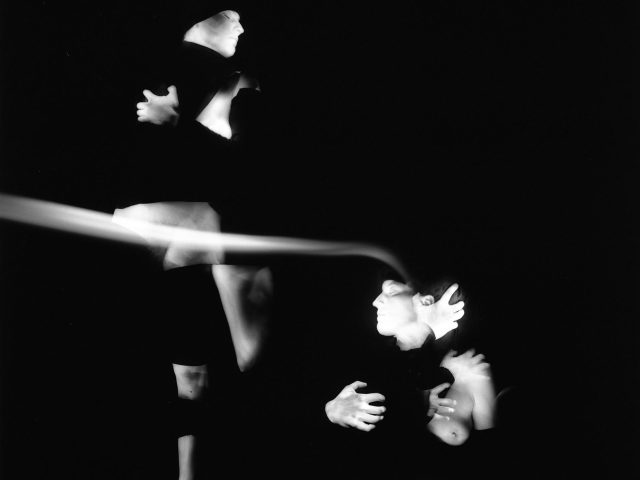 This black and white photograph shows multiple glowing white figures, set against a black background. Their arms are wrapped around themselves and their bodies are blurred with motion.