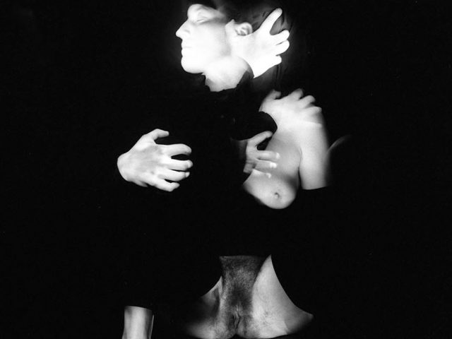 This black and white photograph shows a woman, blurred with motion, with her arms wrapped around herself. Her body glows with light and she wraps her arms around herself.