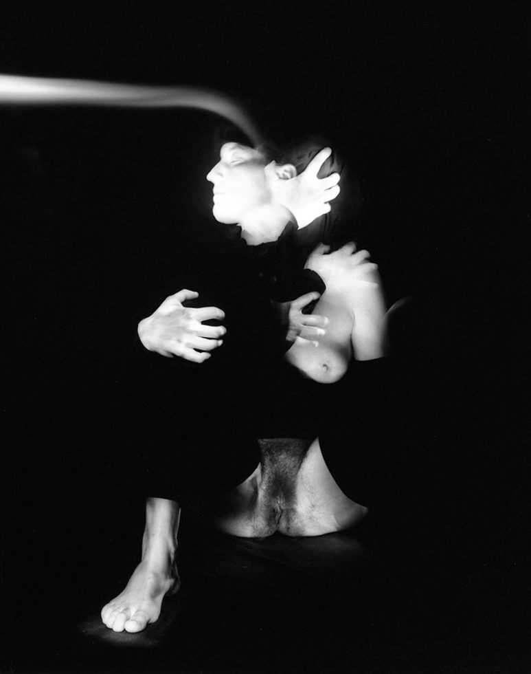 This black and white photograph shows a woman, blurred with motion, with her arms wrapped around herself. Her body glows with light and she wraps her arms around herself.