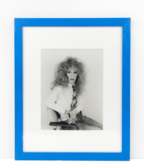 This photograph shows a man wearing a frizzy wig while sitting in a chair.