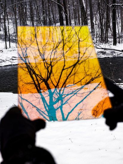 A color photograph of a snowy landscape with the silhouette of hands holding a yellow sheet with the outline of a tree in the foreground.