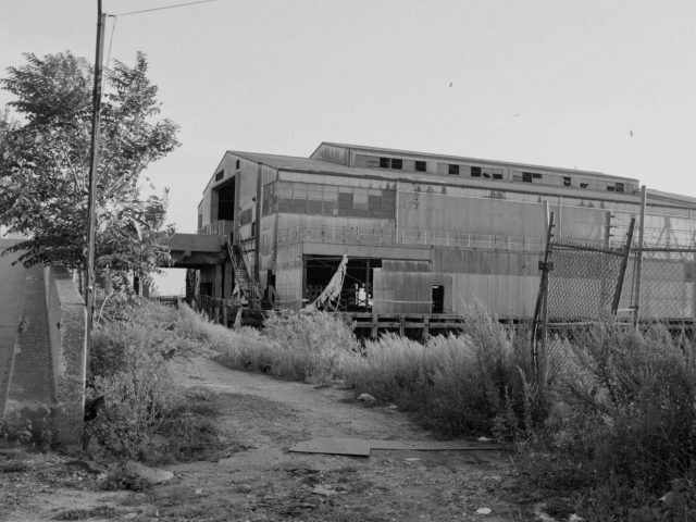 This black and white photograph shows a building sitting at the top of a gravel pathway next to a chain link fence.