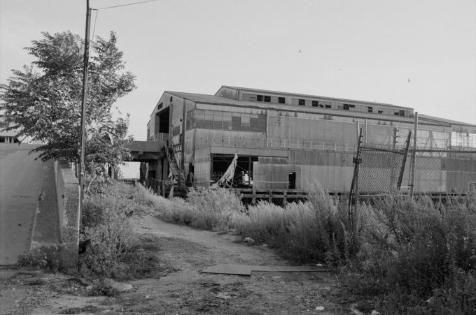 This black and white photograph shows a building sitting at the top of a gravel pathway next to a chain link fence.