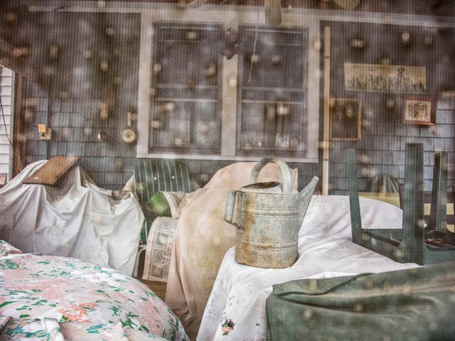 This color photograph shows a screened in porch with some chairs and a watering can.