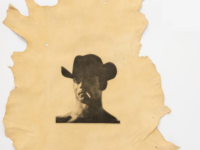 This is a photograph of a man in a cowboy hat, seen from the neck up, printed on an animal hide.