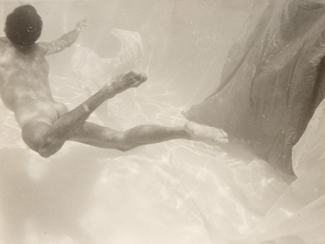 This photograph shows a man swimming underwater with his back to the camera. A piece of fabric floats beside him.