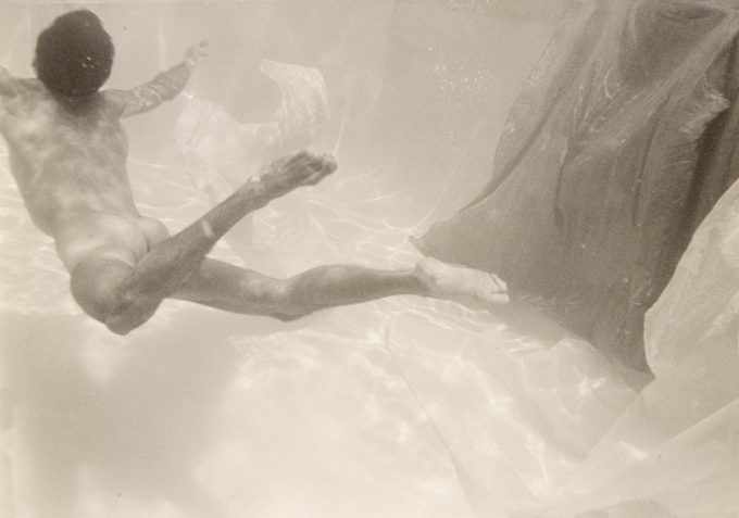 This photograph shows a man swimming underwater with his back to the camera. A piece of fabric floats beside him.