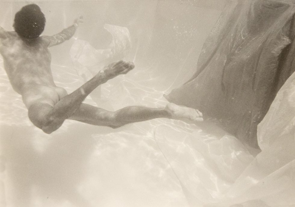 This photograph shows a man swimming underwater with his back to the camera. A piece of fabric floats beside him.