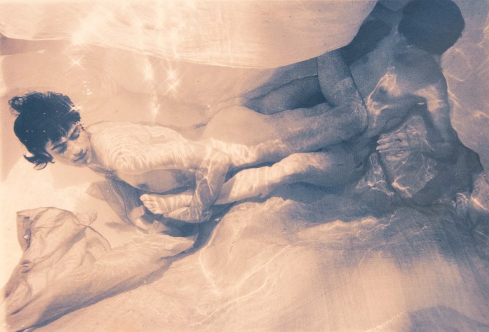This photograph shows a man underwater with his body parallel to the bottom of a pool. Behind him, another man is veiled in floating fabric.