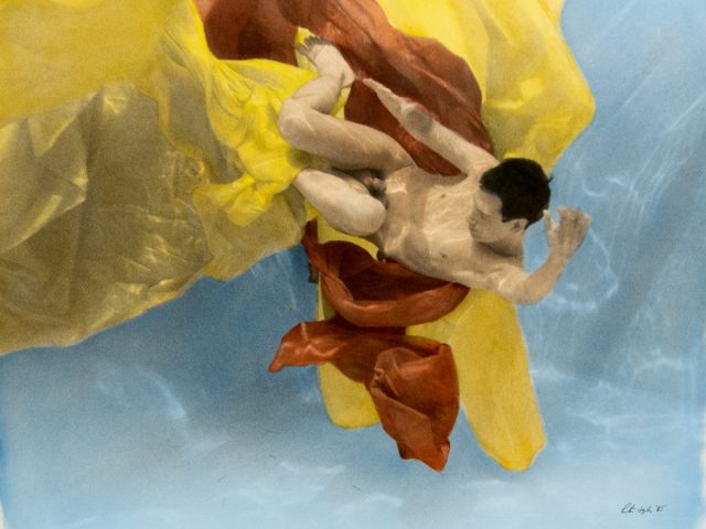 This photograph shows a naked person drifting underwater with billowing yellow and red fabric.