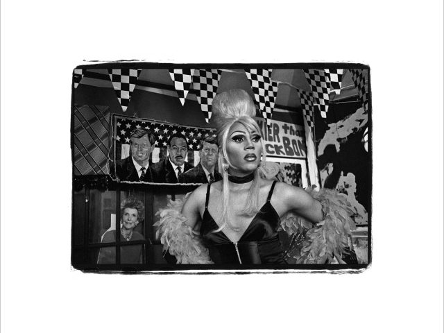 This black and white photograph shows a drag queen staring outside the frame with her hands on her hips. Her hair is very tall and shiny.