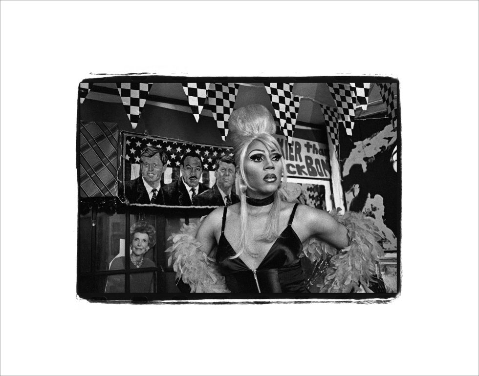 This black and white photograph shows a drag queen staring outside the frame with her hands on her hips. Her hair is very tall and shiny.
