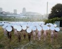 This color image shows a crowd of naked people holding bright white plates above their head in a field.