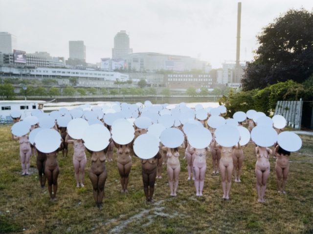 This color image shows a crowd of naked people holding bright white plates above their head in a field.