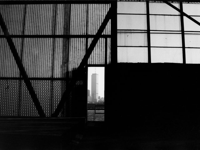 This black and white photographs shows a building seen through an opening in the dark facade of another building under construction.