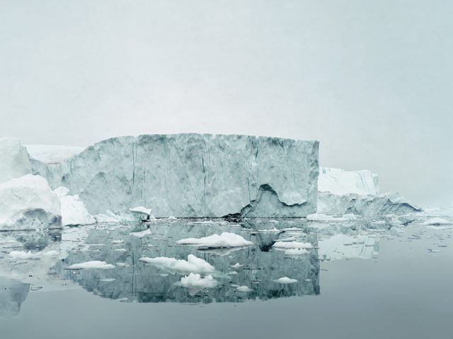 A grayish-blue photo of an iceberg floating with it's reflection below.