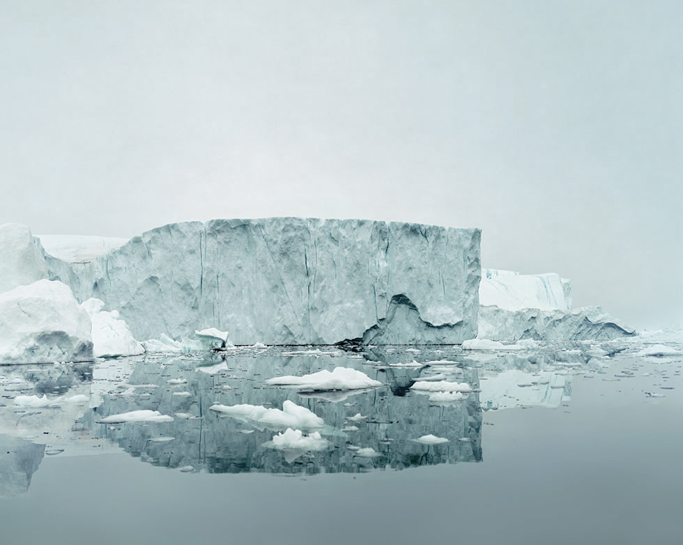 Click to go back. A grayish-blue photo of an iceberg floating with it's reflection below.