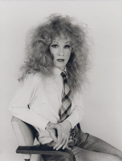 This black and white photograph shows a man wearing a frizzy wig while sitting in a chair.